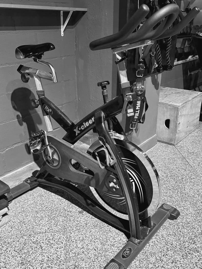 Bicicleta de spinning frente a la pared, con TRX colgados detrás y un cajón de madera al costado.