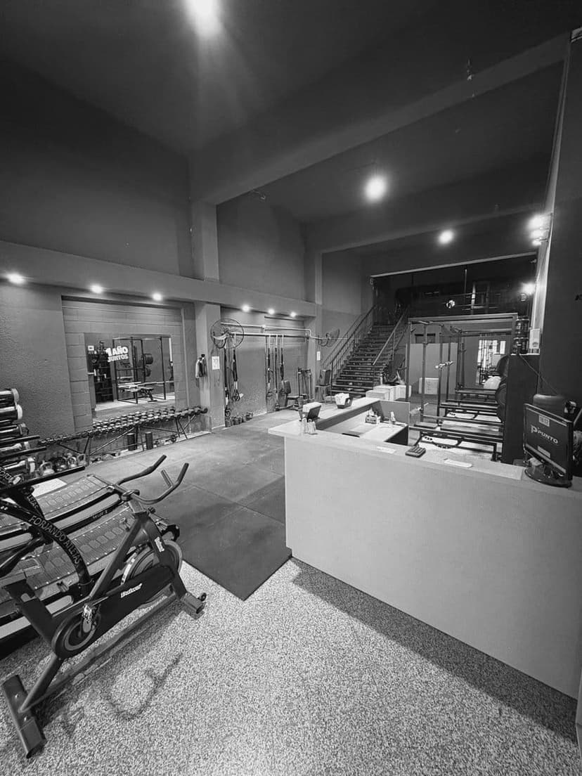 Planta baja desde el mostrador de recepción: bicicleta de spinning y cintas a la izquierda, mancuernas y pelotas contra la pared, TRX colgados al fondo y la escalera a la planta alta.